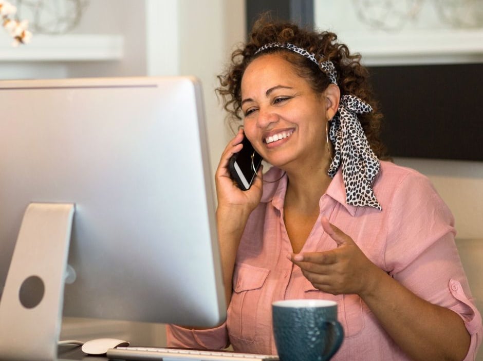 Women wearing a pink shirt, smiling and talking on the phone with a laptop and mug in front of her.