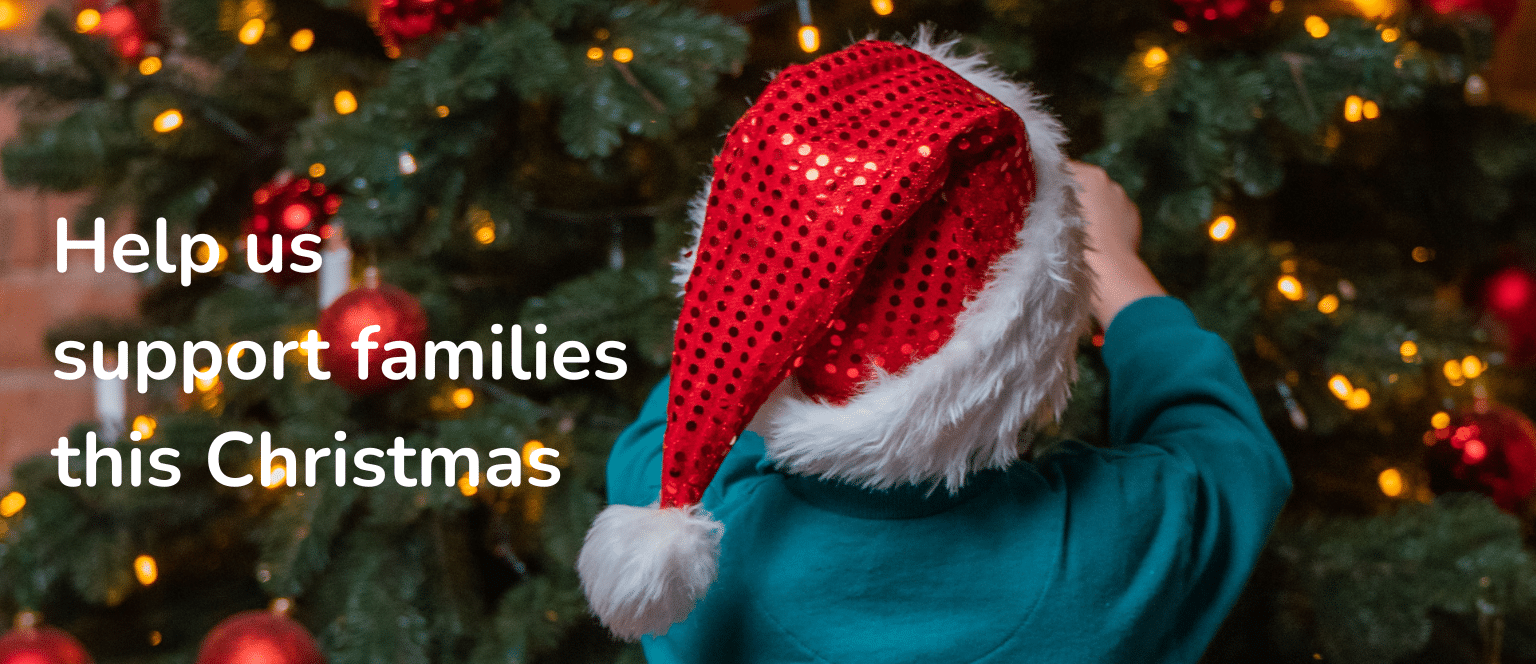 Child in Santa hat hanging decoration on a tree with words 'Help us support families this Christmas'